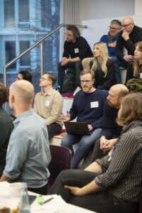Mann mit Laptop auf dem Schoß gestikuliert. Um ihn herum Menschen in einem Konferenzraum.