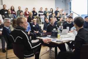 Ein Konferenzraum mit ca 40 Leuten. An einem Tisch spricht ein Mann zu einer Frau.