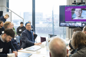Mann während einer Präsentation in einem Konferenzraum. Rechts oben sieht man seine Präsentation auf einem Monitor.