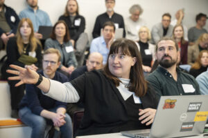 Eine Frau gestikuliert während einer Präsentation. Im Hintergrund: Publikum in einem Konferenzraum.