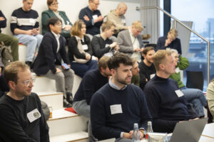 Leute auf Plätzen in einem Konferenzraum. Im Vordergrund ein Mann, der von seinem Laptop aus präsentiert.