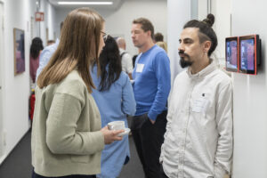 A woman and a man talking in a corridor, with more people in the background.