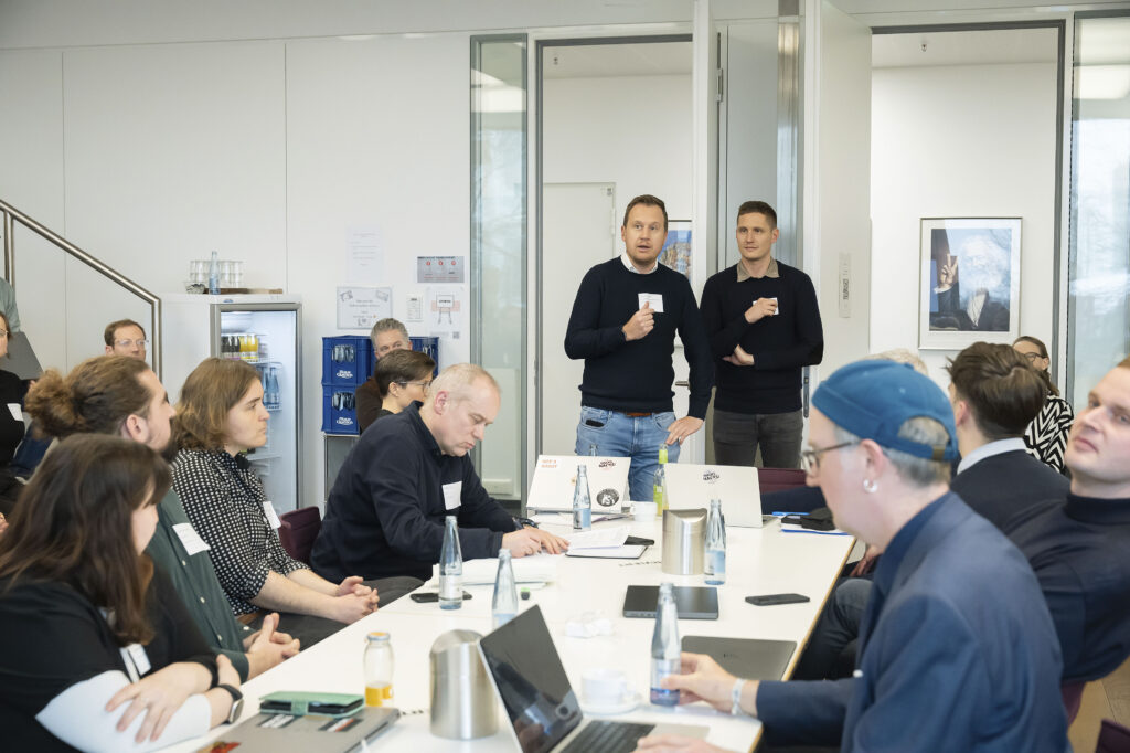 Zwei Männer sprechen stehend in einem Konferenzraum. Das Publikum sitzt an einem Tisch.