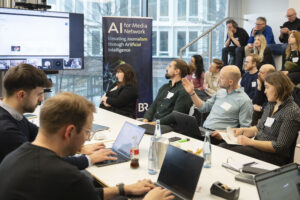 Ein Mann während einer Präsentation. Im Hintergrund Publikum. Im Vordergrund: Menschen