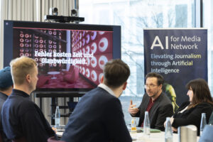 Ein Mann während seines Vortrags. Links ein Monitor mit seiner Präsentation. Hinter ihm ein Roll-Up des AI for Media Network.