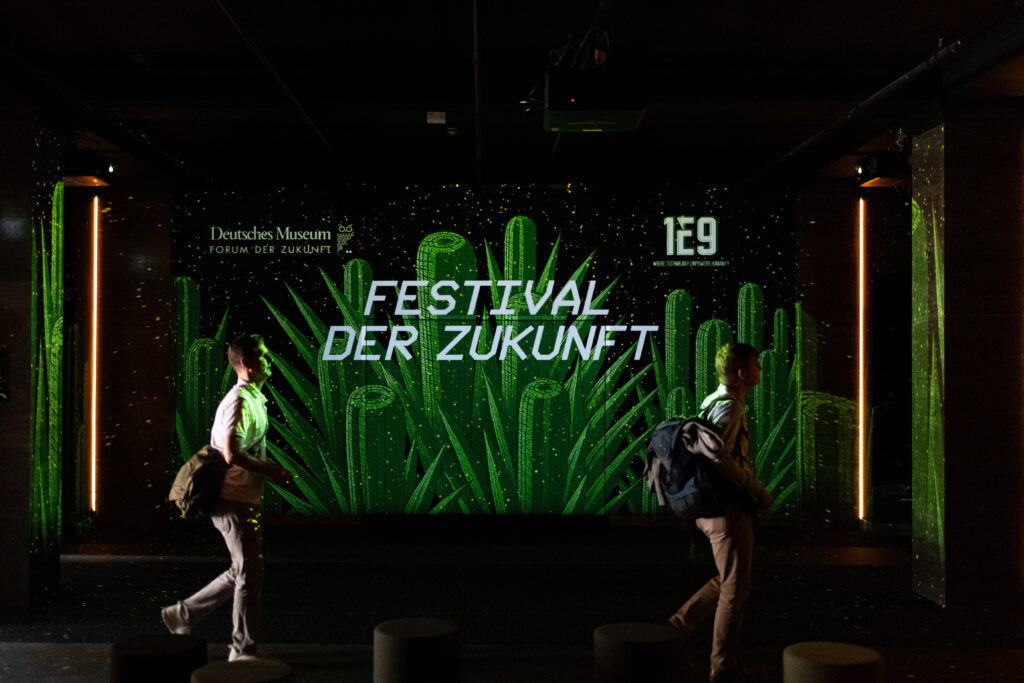Zwei Menschen passieren eine Leinwand mit dem Schriftzug "Festival der Zukunft"